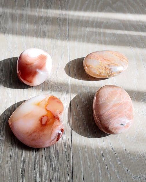 Four polished peach and cream agate and carnelian crystal stones arranged on a light gray wood plank floor, illuminated by natural sunlight with soft shadows.