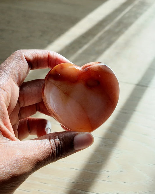 A hand holding a polished heart-shaped agate crystal against a sunlit wooden floor with soft window light and curtains in the background.