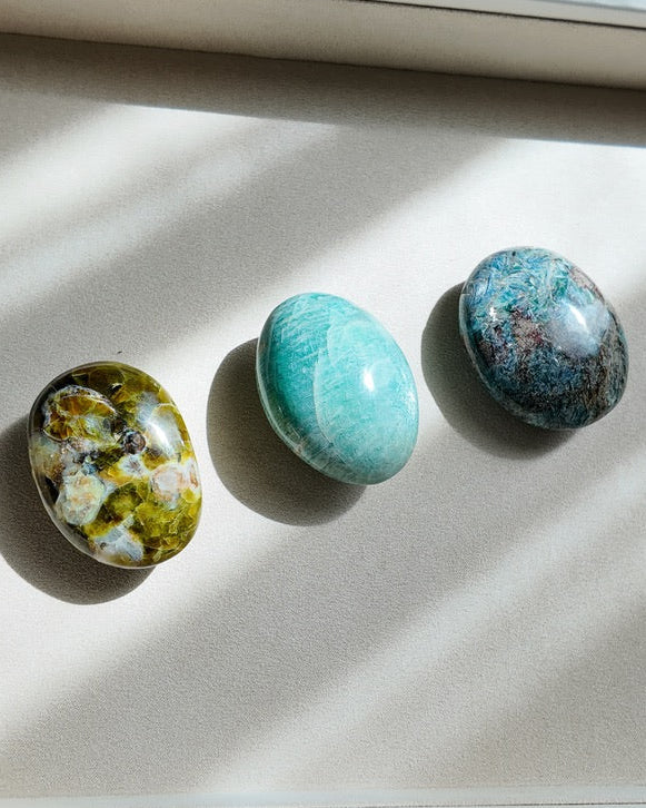 Three polished stone crystals in green, turquoise, and blue hues arranged on a light beige tray with natural sunlight casting shadows.
