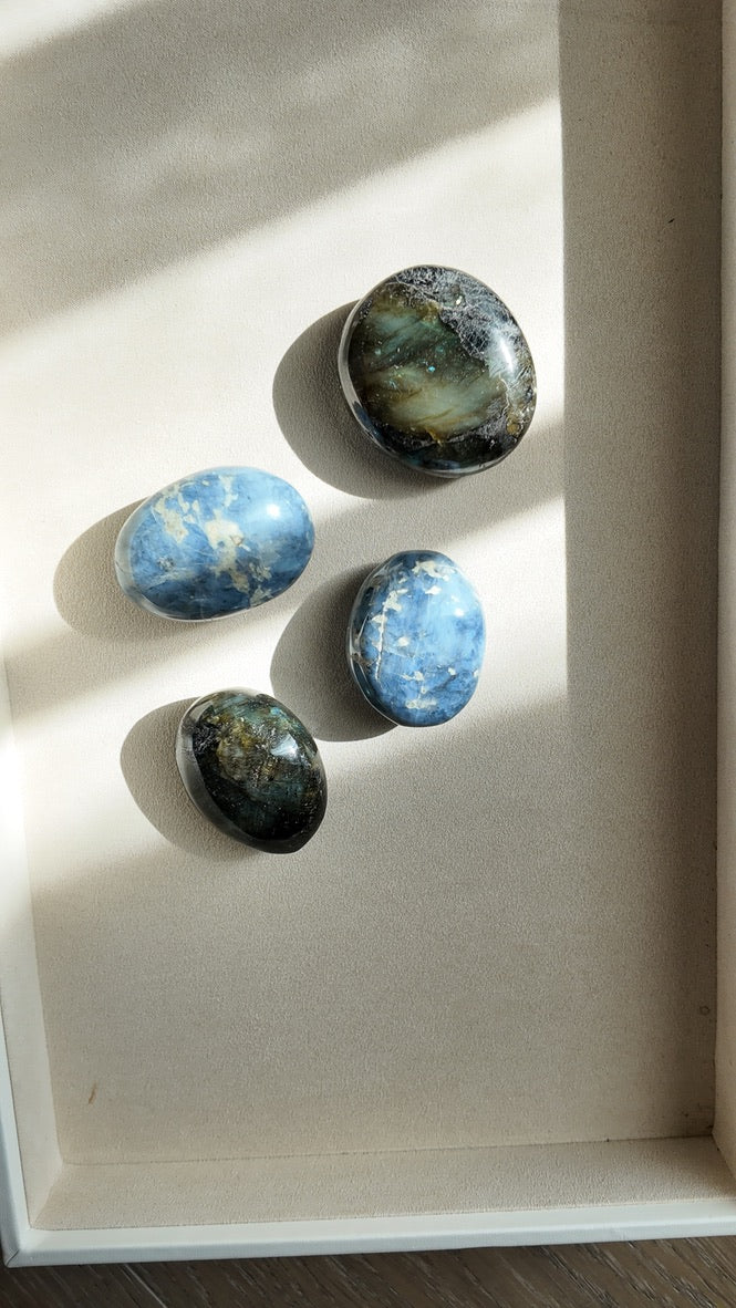 Four polished blue stone pendants with natural dark and light marbling, arranged on a beige textured surface with soft window light casting shadows.