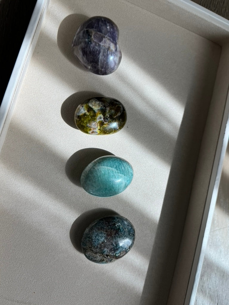 Four polished tumbled gemstones arranged vertically in a white tray on a beige surface, lit by natural sunlight with soft shadows. The stones include purple, green-and-gold marbled, turquoise-blue, and dark blue-gray marbled varieties.