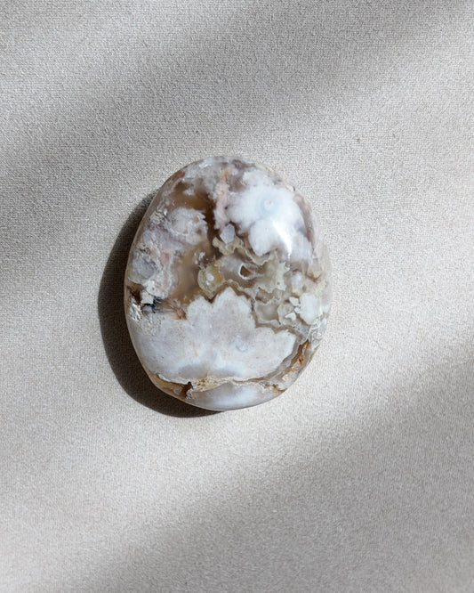 A polished natural stone cabochon with a marbled pattern of white, beige, and brown, resting on a textured beige surface under soft diagonal light. Flower Agate